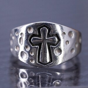 May include: A silver ring with a textured band and a raised cross design.