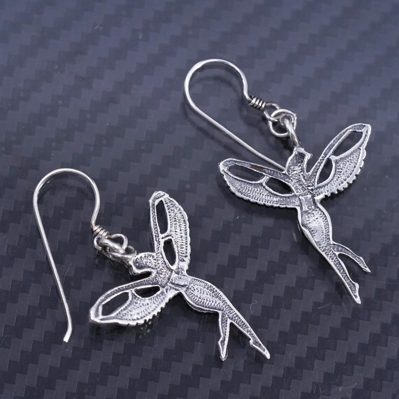 K&ouml;nnte beinhalten: Ein Paar silberne Ohrringe mit einem detaillierten Design einer Fee mit Fl&uuml;geln. Die Ohrringe sind aus Sterlingsilber gefertigt und haben einen zarten, femininen Look.