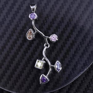 Puede incluir: Un colgante plateado con un diseño en forma de vid, adornado con varias piedras preciosas de colores. El colgante presenta una mezcla de formas, incluyendo piedras redondas, en forma de lágrima, cuadradas y triangulares en tonos púrpura, amarillo y marrón. El colgante está sobre un fondo oscuro.
