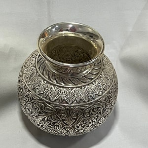 May include: A silver vase with intricate floral designs etched into the surface. The vase has a round body and a wide mouth.