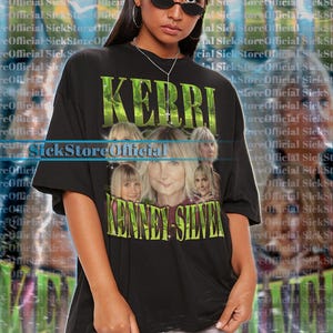 May include: Black unisex t-shirt featuring a graphic of Kerri Kenney-Silver with the text "KERRI" and "KENNEY-SILVER" in green. The shirt is oversized and has short sleeves.