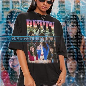 Könnte beinhalten: Schwarzes, übergroßes T-Shirt mit dem Wort "BETTY" in einer farbenfrohen Retro-Schriftart. Das Shirt zeigt eine Grafik einer Gruppe von Personen. Der Text "SickStoreOfficial" ist auf dem Shirt aufgedruckt.
