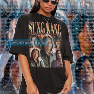 May include: Black unisex t-shirt featuring the name "SUNG KANG" in gold lettering above a graphic of several people. The model is wearing the shirt and sunglasses.