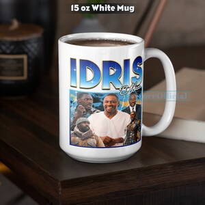 Puede incluir: Taza de cer&aacute;mica blanca con un gr&aacute;fico azul y dorado de Idris Elba y el texto "Idris Elba" en la parte delantera. La taza est&aacute; llena de caf&eacute; y est&aacute; sobre una mesa de madera marr&oacute;n oscura.