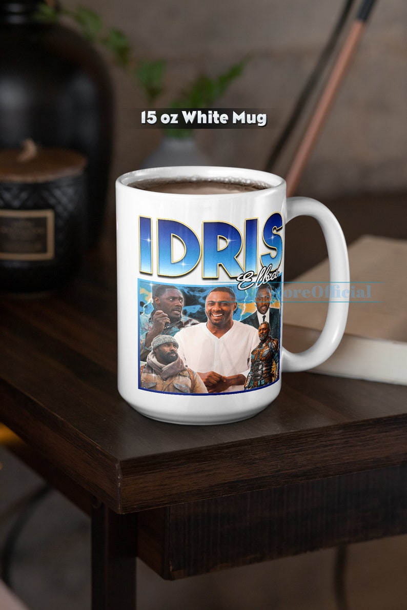 Puede incluir: Taza de cer&aacute;mica blanca con un gr&aacute;fico azul y dorado de Idris Elba y el texto "Idris Elba" en la parte delantera. La taza est&aacute; llena de caf&eacute; y est&aacute; sobre una mesa de madera marr&oacute;n oscura.