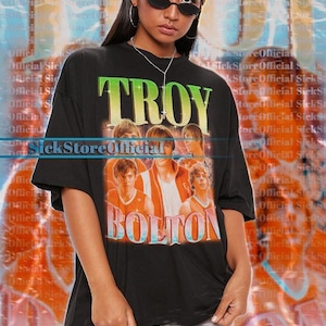 May include: Black oversized t-shirt featuring the text "TROY BOLTON" in a retro style with a photo of a group of young men. The shirt is worn by a person wearing sunglasses.