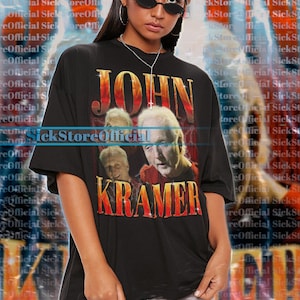 Könnte beinhalten: Schwarzes Unisex-T-Shirt mit dem Text "JOHN KRAMER" in Orange und Gelb. Das Shirt hat eine Grafik von drei Männergesichtern. Das Model trägt eine Sonnenbrille und weiße Shorts. Das Shirt ist übergroß.