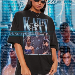 May include: Black unisex t-shirt with a blue graphic of "Katy O'Brian" and a photo collage. The top has short sleeves and a crew neck. The model wears a 2XL shirt. For an oversized fit, choose +1 or +2 sizes.