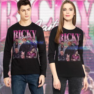 May include: Black long-sleeve shirts featuring a graphic of Ricky Martin with the text "Ricky Martin" in pink and white. The shirts have a crew neck and are worn by a man and a woman.