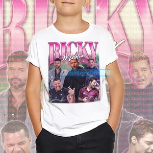 May include: White t-shirt featuring a retro design with the text "Ricky Martin" in pink, along with multiple images of the singer. The design has a vintage aesthetic with a faded background.