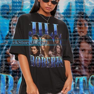 May include: Black oversized t-shirt featuring a graphic of Julia Roberts and the text "JULIA ROBERTS" in a blue, stylized font. The shirt is a unisex style and is worn by a person wearing sunglasses.