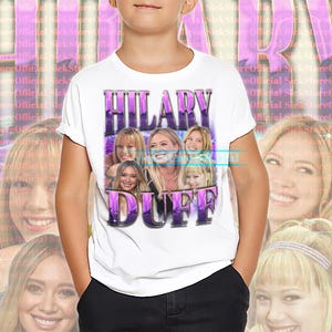 May include: White t-shirt featuring a graphic of Hilary Duff in a purple and white design. The shirt has the text "HILARY DUFF" in a retro font. The background has a repeating pattern of the text "SickStoreOfficial".