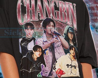 May include: Black unisex t-shirt featuring a graphic of a musical artist with the name "CHANGBIN" in a distressed, vintage-style font. The shirt has a crew neck and short sleeves. The image includes multiple figures in various poses.