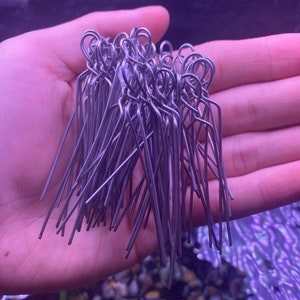 May include: A collection of silver-colored metal pins, each with a looped top and a straight, pointed bottom. The pins are held together in a hand, creating a dense bundle of wire. The background is a blurred, aquatic-themed scene.