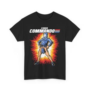 Puede incluir: Camiseta negra con un gráfico de un personaje de dibujos animados de Cobra Commando con un mono azul y un casco, de pie en una explosión roja y amarilla. El texto "COBRA COMMANDO" está por encima del personaje.
