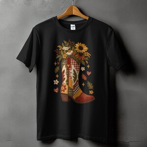 May include: A black short-sleeved t-shirt featuring a graphic of a cowboy boot filled with a bouquet of sunflowers and autumn leaves. The boot has a patchwork design with red, yellow, and green patterns, surrounded by small hearts, stars, and flowers.