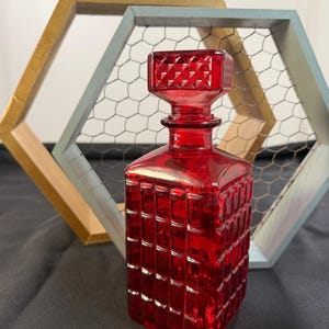 May include: A ruby red glass decanter with a geometric pattern and a square stopper. The decanter is rectangular and sits on a dark surface. In the background are two hexagonal wooden frames with chicken wire.
