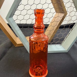 May include: An orange glass bottle with a textured surface and a round base. The bottle has a narrow neck and a decorative design. The background includes a decorative wooden frame with chicken wire.