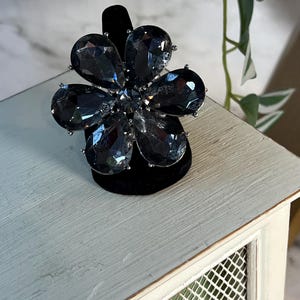 May include: A black flower-shaped ring with large, faceted, dark gray stones. The ring is on a black velvet display stand.