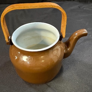 May include: A brown enamel teapot with a wooden handle. The teapot has a spout and a light blue interior. The handle is made of light brown wood and is attached to the pot with dark brown metal brackets.