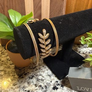 May include: Gold-toned bracelets displayed on a black velvet stand. One bracelet features a leaf design adorned with small, clear crystals. The other bracelets have a textured, rope-like pattern. A small ring with a similar design is also visible.