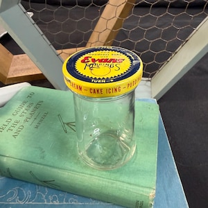 May include: A clear glass jar with a yellow and blue lid labeled "Evans Toppings" sits atop a vintage green book. The lid features text that reads "Cream - Cake Icing - Pudding". The jar is empty.