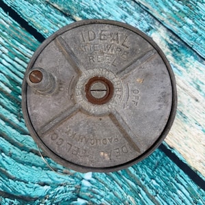 May include: A vintage Ideal Tie Wire Reel, made of weathered metal. The reel has the text "IDEAL TIE WIRE REEL" and "PADUCAH, KY" embossed on its surface. The reel is circular with a central spindle and a side knob.