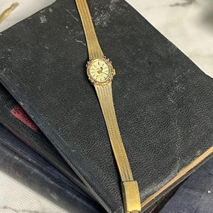 May include: A gold-toned wristwatch with a white face and a gold band. The watch has a small, round face with black Roman numeral hour markers and a small second hand. The band is a delicate, chain-link style. The watch is surrounded by a ring of small, pink stones.