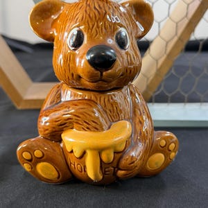 Puede incluir: Olla de miel de cerámica marrón con forma de oso. El oso está sentado y sostiene una olla de miel amarilla con la palabra "HONEY" escrita. El oso tiene ojos negros y nariz negra. Es un artículo de cocina.
