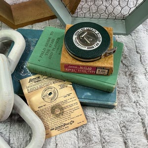 May include: A vintage green and gold Lufkin Ni-Clad Royal Steel-Tape measure sits atop a small box and a green book titled "A Field Guide to the Stars and Planets". A vintage instruction sheet is also visible.