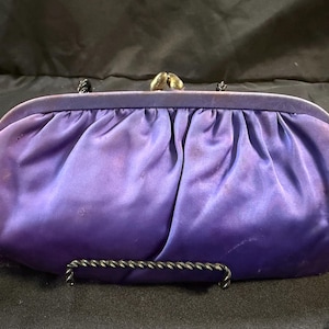 Può includere: Una pochette vintage in raso viola con chiusura e manico in metallo. La borsa ha un design a pieghe e un manico in metallo scuro. Il tessuto di raso presenta qualche scolorimento.