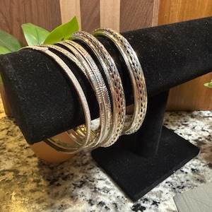 May include: A collection of silver bangles displayed on a black velvet jewelry stand. The bangles have various designs, including textured and openwork patterns. The background includes a wooden surface and greenery.