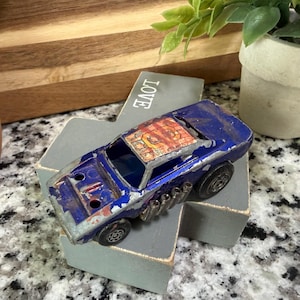 May include: A vintage blue toy car with a distressed finish, featuring a detailed roof design and side pipes. The toy car is resting on a gray block with the word "LOVE" visible. The car's wheels are black.