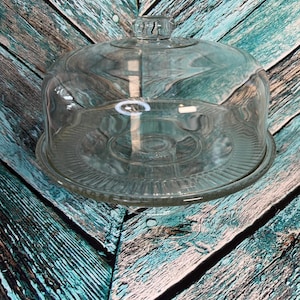 May include: A clear glass cake stand with a domed lid. The stand has a round base with a ridged pattern and a clear glass dome with a small handle on top. The stand is on a blue and brown wooden surface.