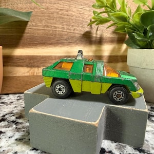 May include: A vintage toy car in green and yellow, with black wheels and orange tinted windows. The toy car is sitting on a gray block, with a wooden background and a potted plant in the background.