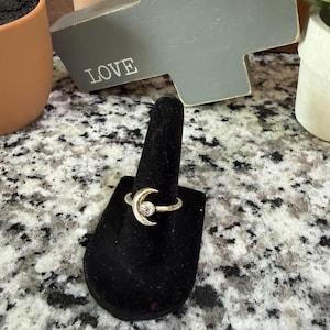 May include: A silver-toned ring featuring a crescent moon design with a clear, round gemstone at its center. The ring is displayed on a black velvet ring holder. The word "LOVE" is visible in the background.