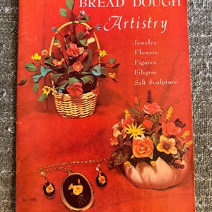 May include: A red instructional booklet titled BREAD DOUGH Artistry featuring text listing Jewelry, Flowers, Figures, Filigree, and Salt Sculpture. The cover displays a woven basket of dough flowers, a floral arrangement in a bowl, and a necklace set.
