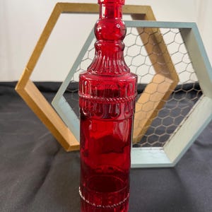 May include: A tall, cylindrical red glass bottle featuring a narrow neck, a cork stopper, and decorative embossed patterns including a rope-like band around the upper body and vertical ridges near the base and neck area.