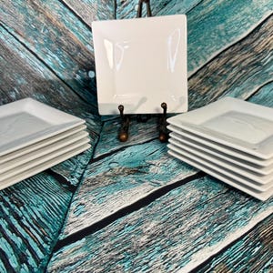 Può includere: Pile di piatti quadrati bianchi. I piatti sono disposti in tre pile, con un piatto esposto su un supporto decorativo. I piatti hanno un design semplice, perfetto per servire antipasti o dessert. Lo sfondo è in legno rustico e invecchiato.