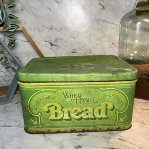 Op de afbeelding: Vintage groene metalen broodtrommel met de woorden "Wheat Heart Bread" in gele letters. De rechthoekige trommel heeft een scharnierend deksel. De woorden "Oven-Baked" en "Enriched" zijn ook zichtbaar.