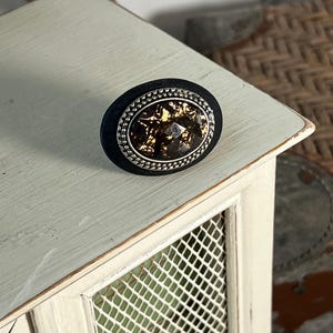 May include: A black oval-shaped knob with a silver rim and a gold-toned, textured center. The knob is sitting on a white surface with a wire mesh background.
