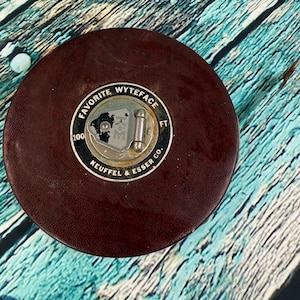 May include: A vintage brown leather Favorite Wyteface measuring tape by Keuffel & Esser Co. The tape measure has a metal face with the text "Favorite Wyteface" and "100 FT". The tape measure is round.