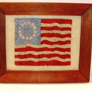 May include: A framed needlepoint artwork depicting the American flag with 13 red and white stripes and a blue canton with 13 white stars in a circle. The frame is made of dark wood.