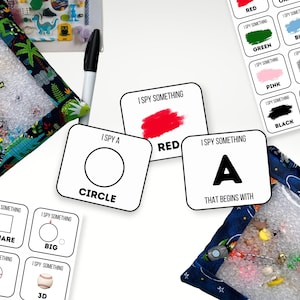 May include: Two sensory bags with clear windows filled with small objects, surrounded by I Spy game cards featuring shapes, colors, and letters. One bag has a dinosaur print, the other a space print. A black marker is included for the activity cards.