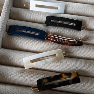 Peut inclure: Collection de barrettes rectangulaires pour cheveux de différentes couleurs et motifs. Les barrettes comprennent du blanc, du noir, bleu marine, écaille de tortue et blanc cassé avec des accents métalliques dorés. Présentées sur un tissu neutre.