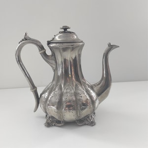 Puede incluir: Una tetera de plata con un diseño ornamentado y un motivo floral en la tapa. La tetera tiene un pico curvo y un asa con un inserto de madera.