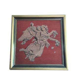 Op de afbeelding: Een ingelijste tapijtkunst met een klassieke scène met figuren, engelen en bloemelementen. Het kunstwerk is voornamelijk in grijstinten en beige, tegen een rode achtergrond. De lijst is goudkleurig.