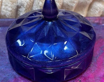 May include: A collection of resin art pieces in shades of blue and purple. The set includes a lidded, faceted jar, a tray, a mushroom-shaped sculpture, and a small square container. The items have a glittery, iridescent finish, creating a shimmering effect.