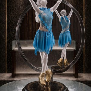 Op de afbeelding: Een beeldhouwwerk van een kunstschaatser in een blauwe jurk met bloemaccenten en gouden schaatsen. De figuur poseert met opgeheven armen, weerspiegeld in een ronde spiegel. Het beeld staat op een zwarte basis met de letters "MNi".