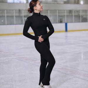 Op de afbeelding: Een zwart kunstschaatskostuum met een hoge kraag en lange mouwen. Het kostuum wordt gedragen met witte schaatsen. De schaatser staat op een ijsbaan met een gele en blauwe rand.